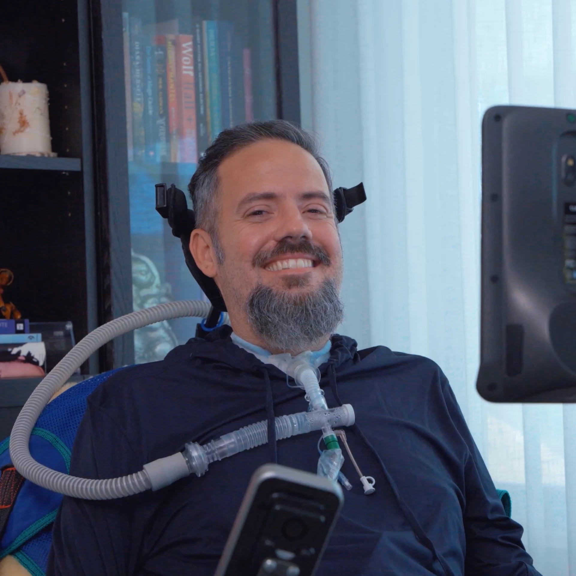 A man with a beard smiling, sitting in a wheelchair with medical equipment, in front of a bookshelf and a window.